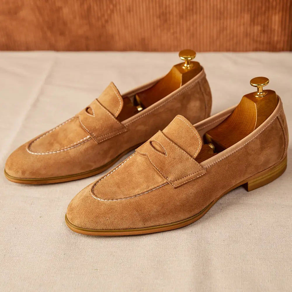 Chaussures de type loafers en suède couleur camel au design classique, avec surpiqûres contrastantes blanches et des semelles en cuir brun clair.