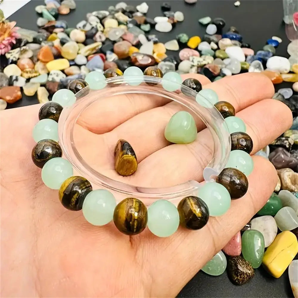 A hand holds a delicate natural tiger eye and green aventurine beaded bracelet featuring alternating smooth light green jade-like beads and polished golden-brown tiger eye stones.
