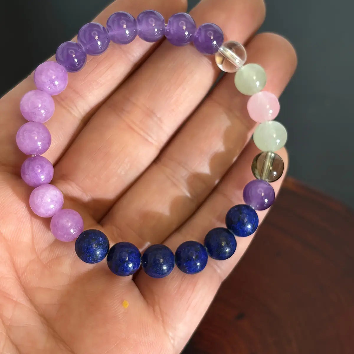 A hand holds a multicolor gradient fluorite beaded bracelet featuring smooth round stones in shades of purple pink green and deep blue with a crystal accent.