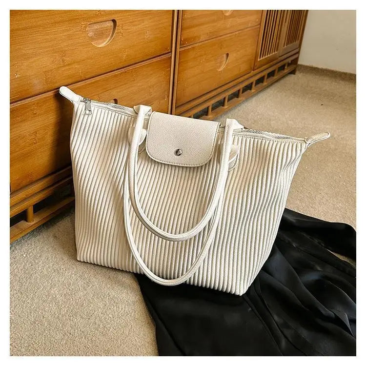 A cream-colored ribbed leather minimalist pleated shoulder tote bag with a matching flap closure and silver-toned hardware.