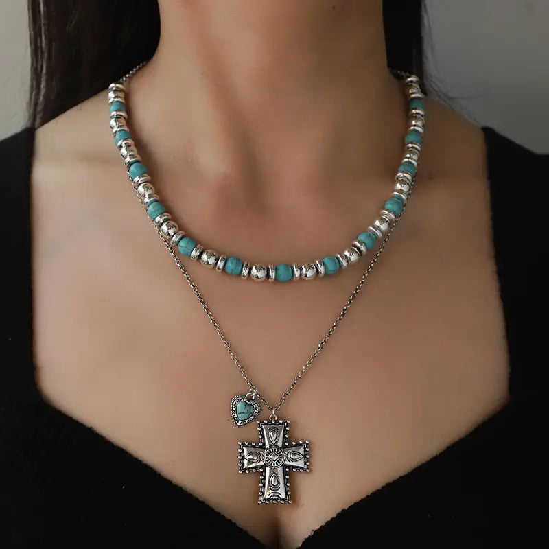 Eine Lagenhalskette aus dem Schmuckset „Turquoise Heart and Sunburst Silver Jewelry Set“ mit türkisfarbenen Perlen und silberfarbenem Metall, mit einem markanten Kreuzanhänger, der mit aufwendigen Details verziert ist.