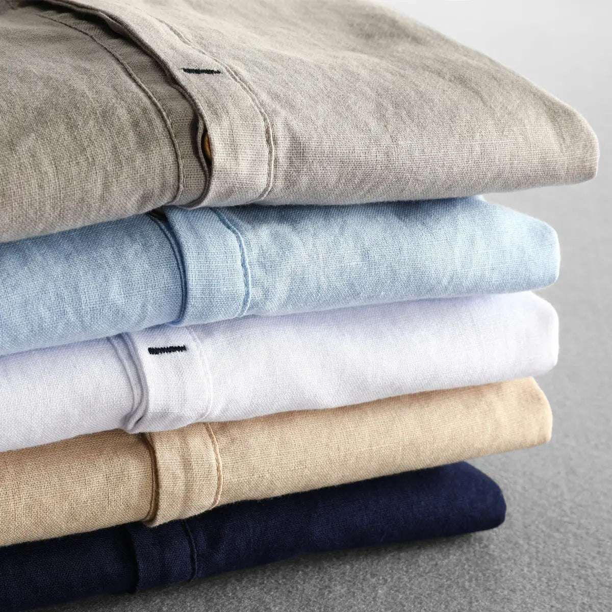 Stack of neatly folded men’s loose linen pants in neutral tones including beige light blue white tan and navy accompanying a loose linen mandarin collar long sleeve shirt.
