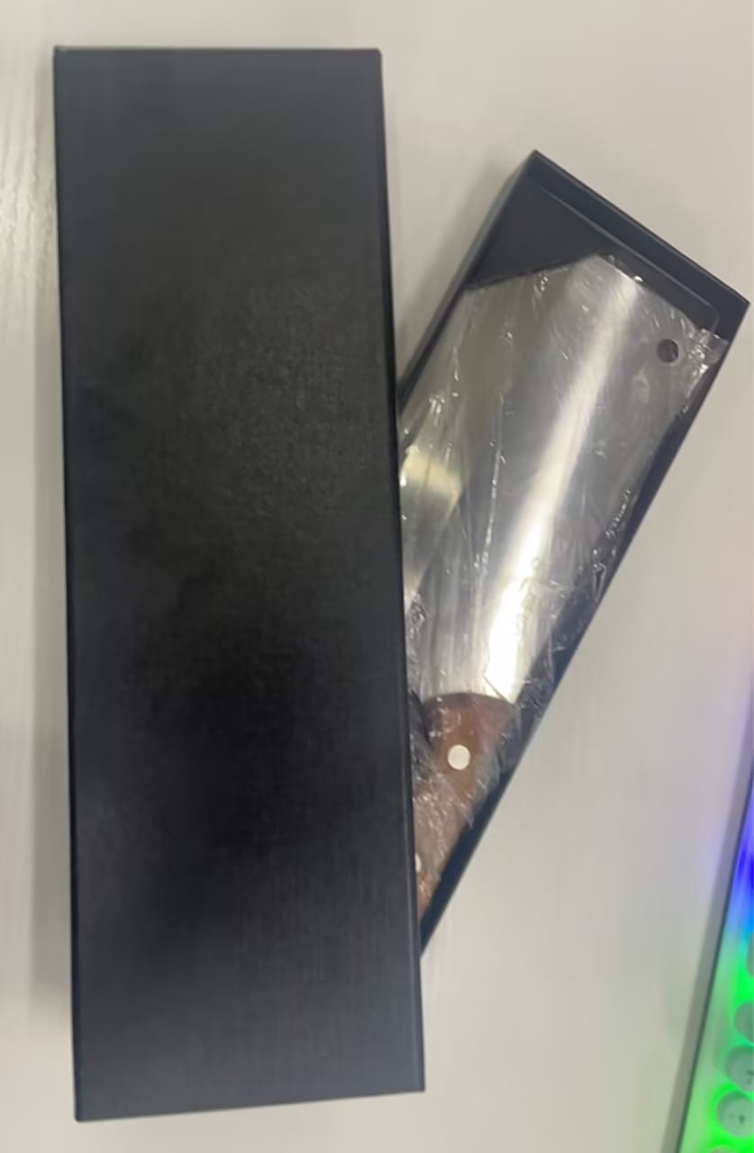 Une hache/couteau de chef en acier inoxydable avec une lame polie et un manche en bois foncé reposant à l’intérieur d’une boîte cadeau noire.