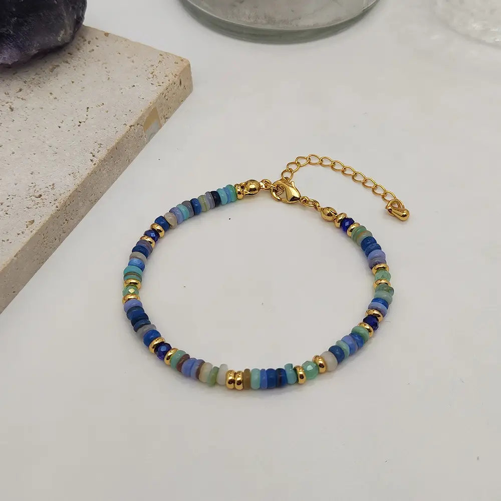 A delicate handwoven rainbow shell beaded bracelet with blue green and purple stones featuring gold-toned metal accents and a gold clasp.