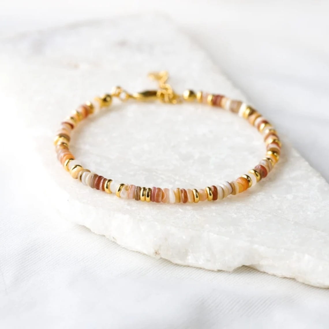 A handwoven rainbow shell beaded bracelet featuring alternating peachy-orange and creamy white cylindrical beads with gold-toned metallic spacers and a gold clasp.