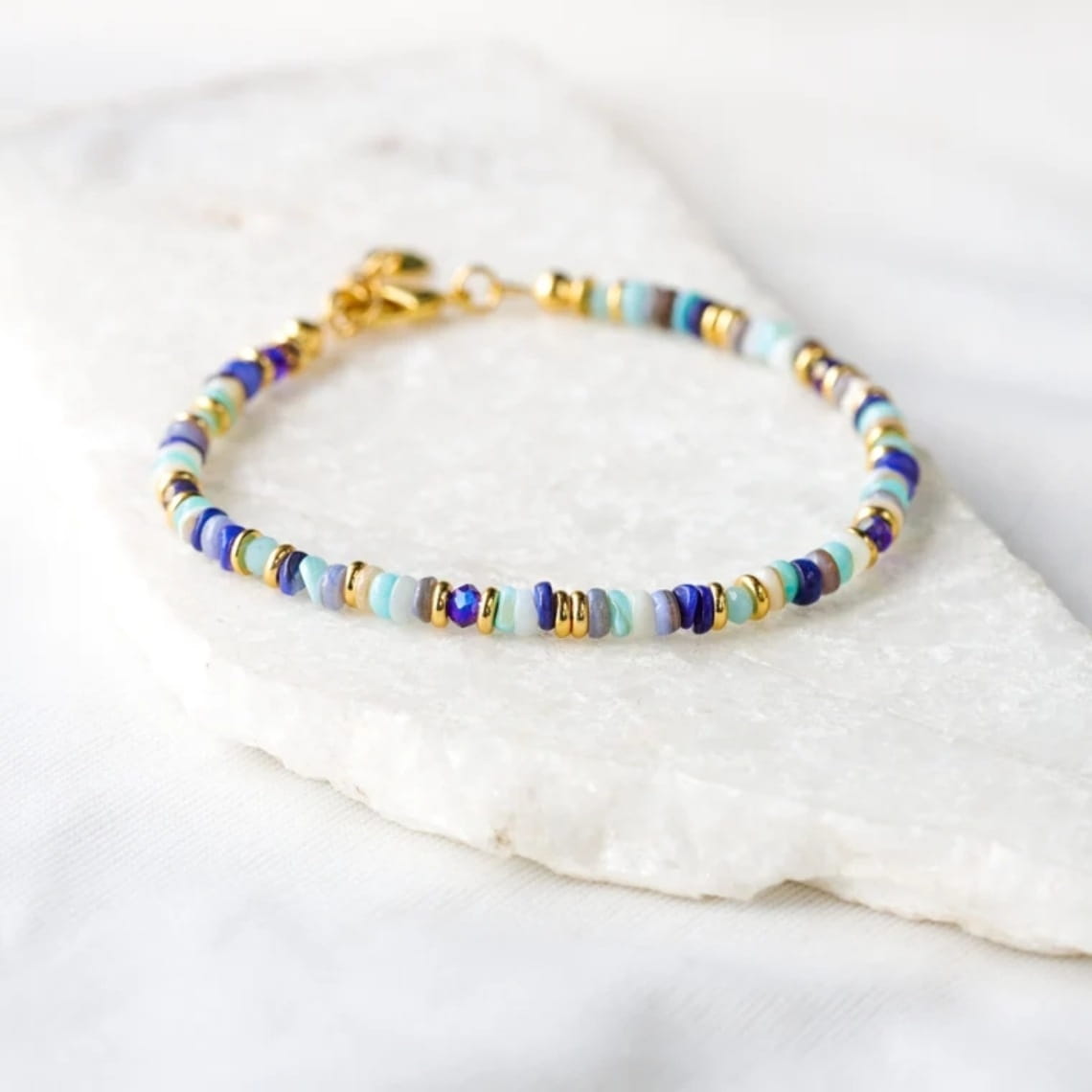 A handwoven rainbow shell beaded bracelet with gold clasp featuring delicate blue teal and gold-toned beads and a small heart-shaped accent.