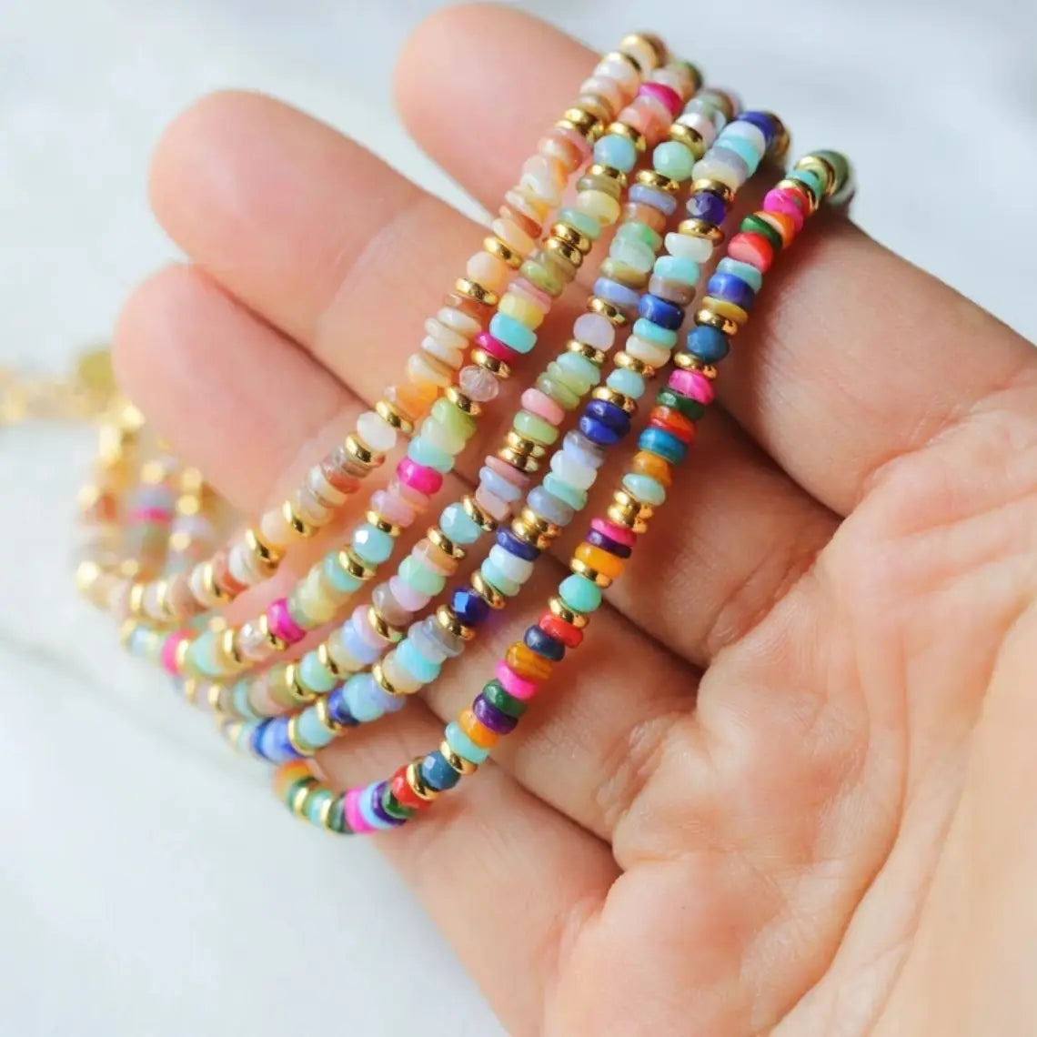 A hand holds colorful handwoven rainbow shell beaded bracelets featuring multicolored gemstone-like beads with gold-toned metal spacers and gold clasps.