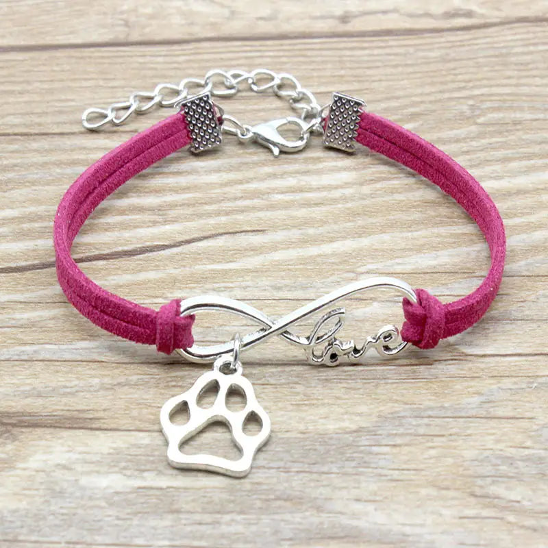 A pink suede hand-woven bracelet featuring a silver infinity charm engraved with love and a dangling paw print pendant secured by a silver clasp.