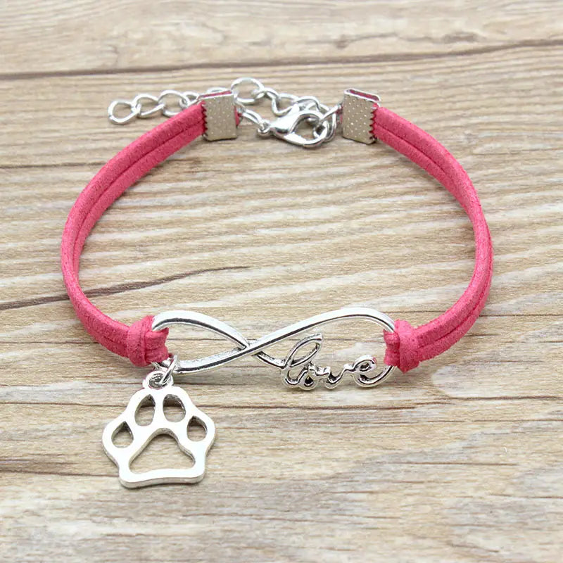 A pink suede hand-woven bracelet featuring a silver infinity charm engraved with love and a dangling paw print pendant designed for pet lovers.