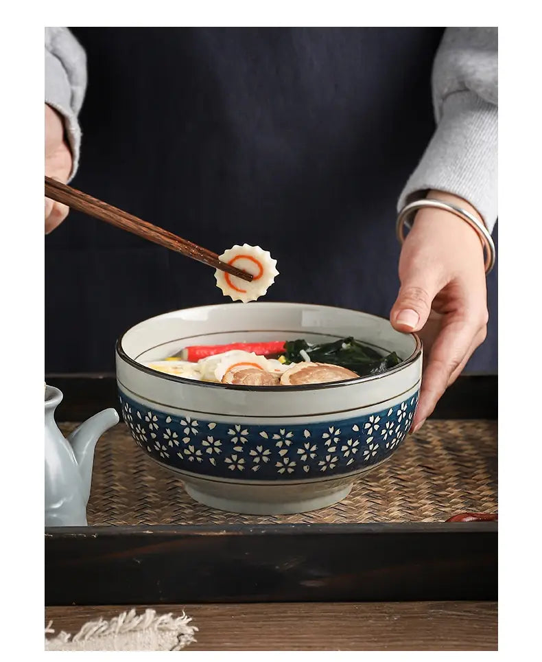 A ceramic noodle bowl 7 inch cherry blossom soup bowl featuring a navy blue band with white floral patterns and a matching rim, holding steaming soup.