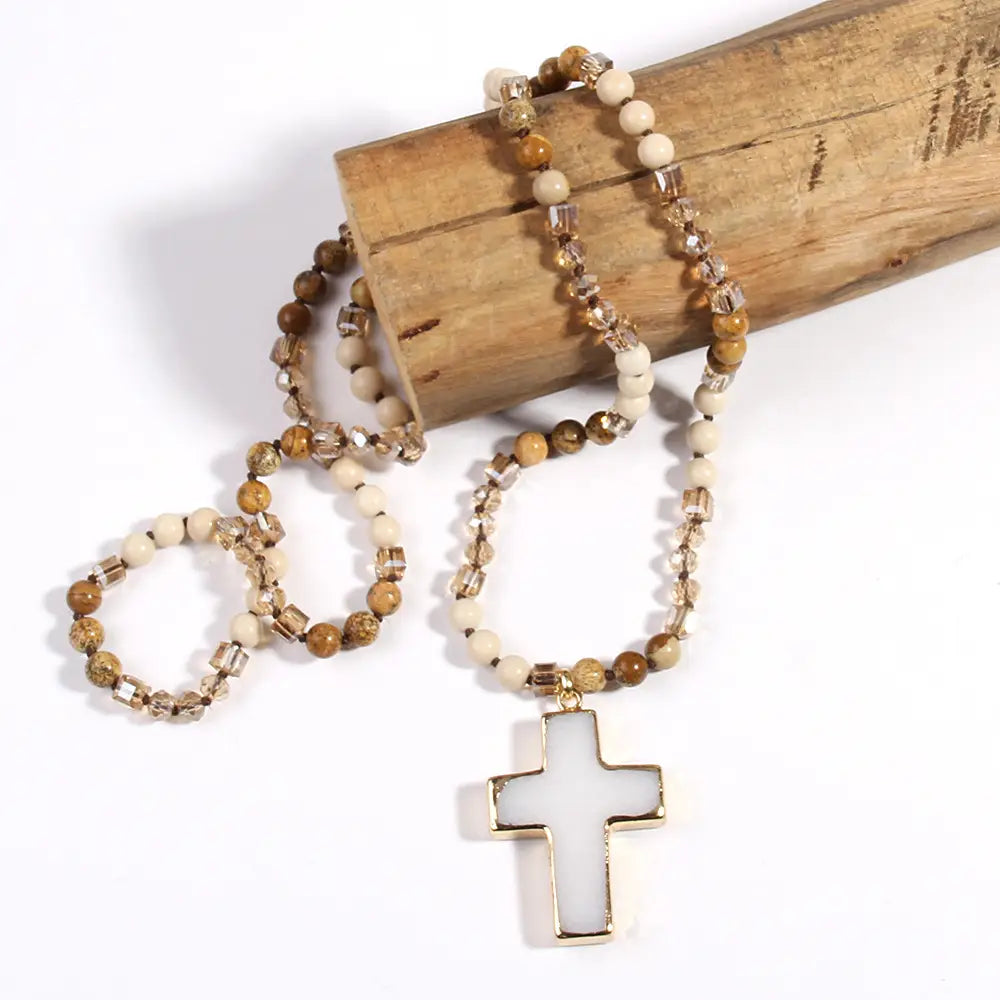 A delicate bohemian natural stone and glass beaded cross necklace with a white cross pendant featuring alternating earthy brown and cream-colored beads with clear crystal accents.