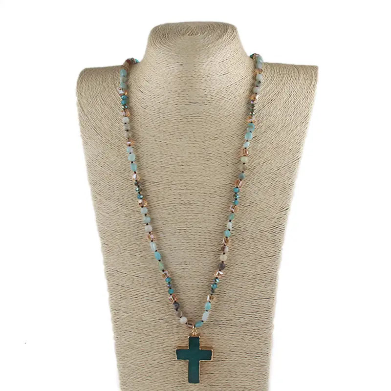 A bohemian natural stone and glass long beaded necklace with turquoise and white stones featuring a dark green cross pendant.