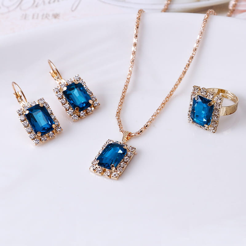 A set of elegant gold-toned earrings necklace and ring featuring a central rectangular blue gemstone surrounded by smaller clear crystals in a three piece jewelry set.