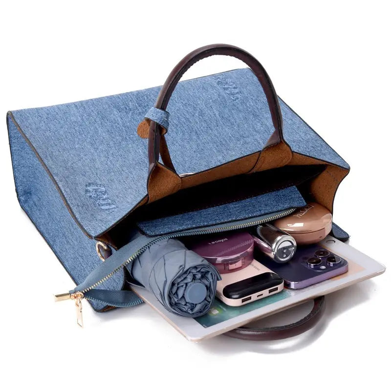 Denim blue handbag with brown leather handles and interior lining featuring a zippered compartment holding personal items from the denim and leather three piece multi compartment handbag set.