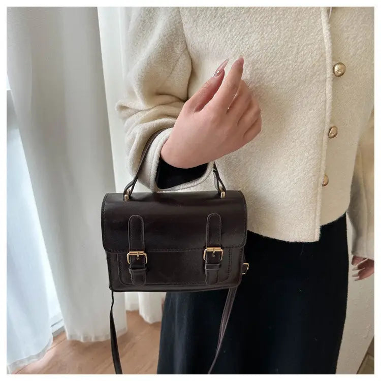 Sacoche vintage en effet cuir synthétique brun foncé, avec des boucles en ton or et une sangle amovible présentée à l’avant.