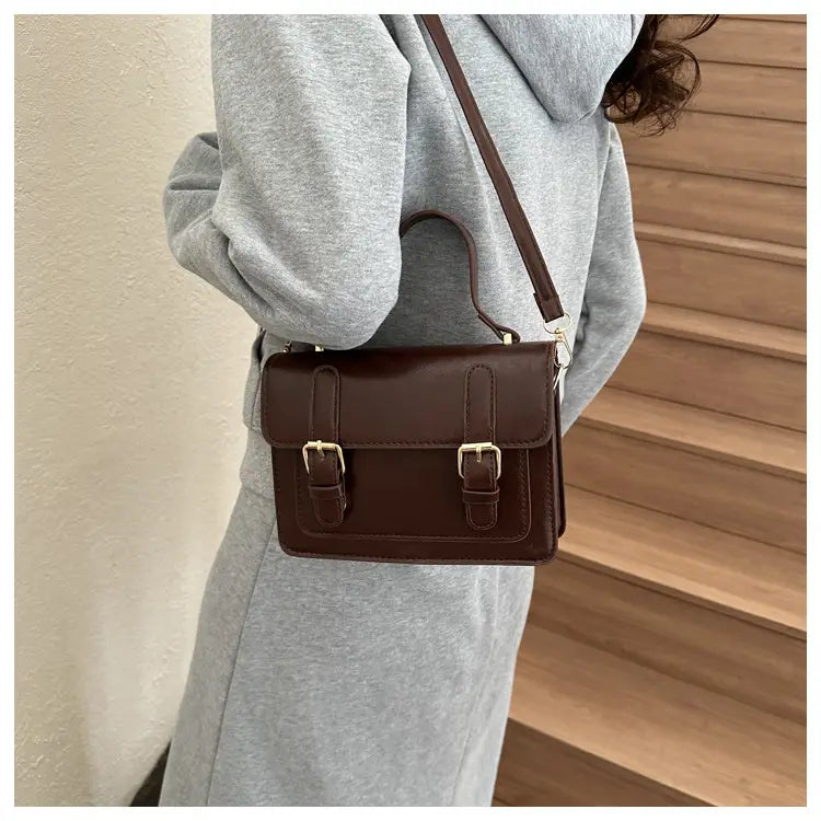 Sacoche vintage en cuir brun à effet cuir synthétique, avec deux fermetures à boucles en ton or et une bandoulière assortie.