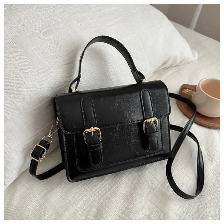 Sacoche vintage en effet cuir synthétique noire, avec fermetures à boucles en ton or et une bandoulière amovible.