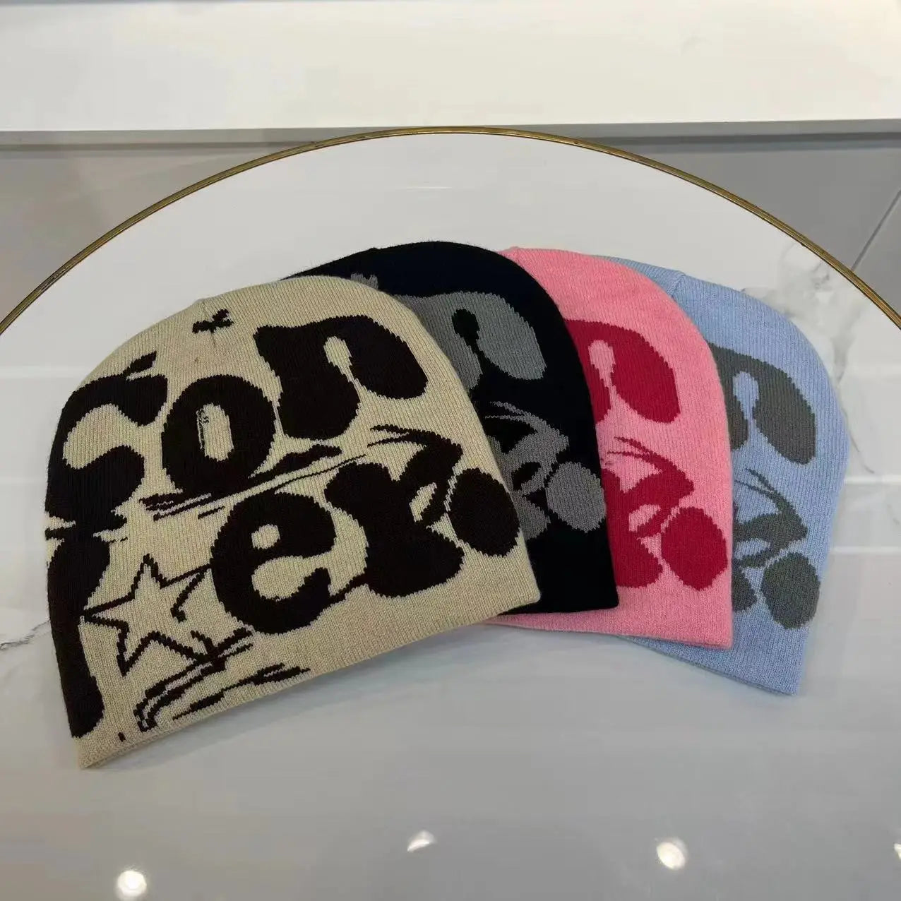 A collection of knitted letter jacquard beanie warm winter hats in beige black pink and light blue featuring bold graphic lettering with a star motif.