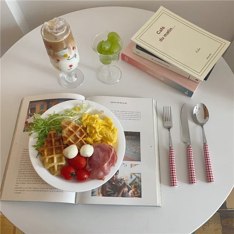 White plate with waffle scrambled eggs cherry tomatoes mozzarella balls and ham served with a checkered stainless steel cutlery tableware set.