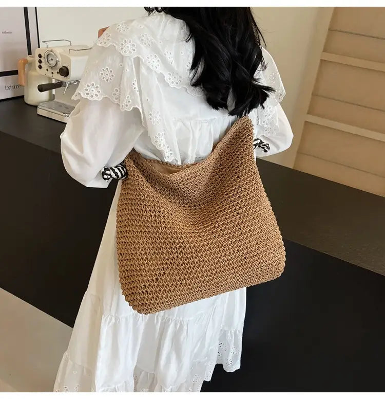 A tan hand-crocheted straw tote bag with a soft textured weave and wide shoulder strap featuring striped bow handles.