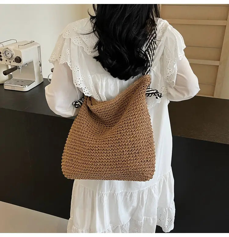 A tan hand-crocheted straw tote bag with a soft textured weave and striped bow handles featuring a cozy slouchy silhouette.