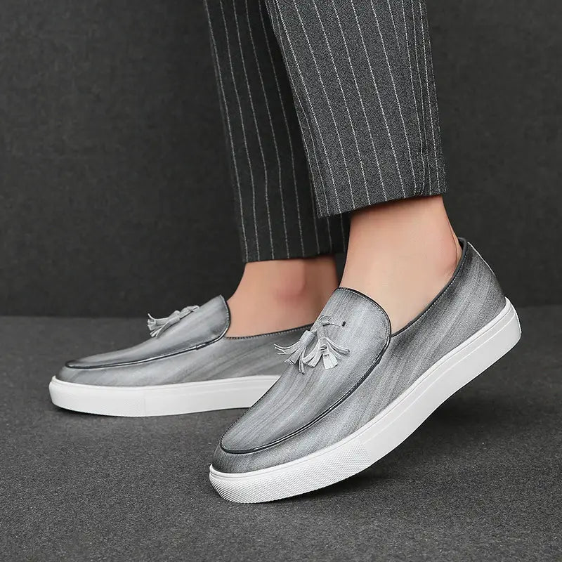 Mocassins en cuir gris pour homme, à enfiler, avec semelles en caoutchouc blanc et ornements à glands sur l’empeigne.