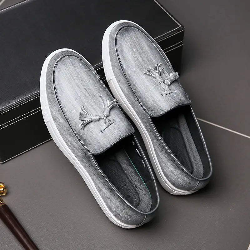 Mocassins à enfiler en daim gris pour homme, avec semelles en caoutchouc blanc et ornements à glands sur l’empeigne, contrastant avec les mocassins à glands en cuir brun à semelle blanche, intitulé.