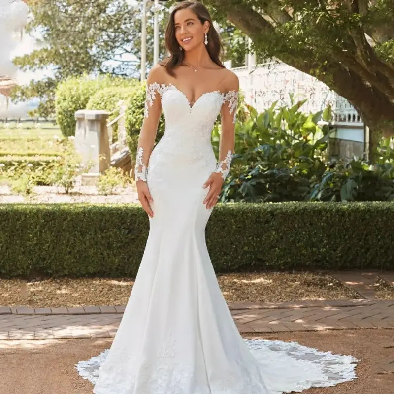 Une robe de mariée sirène en dentelle à épaules dénudées blanche, avec des détails de dentelle complexes sur les manches et le corsage, présentant une silhouette ajustée qui s’évase en une traîne fluide.