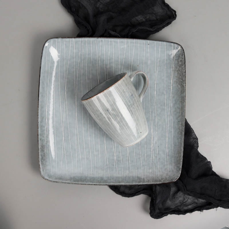 a light blue ceramic mug with vertical white streaks and a rustic handcrafted finish resting on a matching square plate from the ceramic dinnerware set retro ribbed plates and bowls