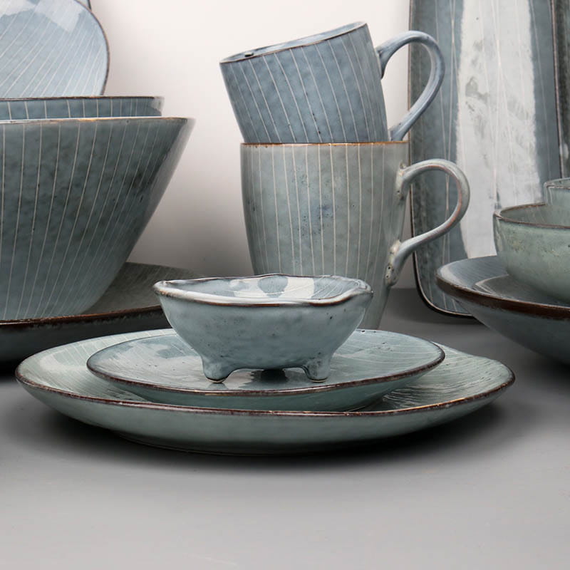A rustic light blue ceramic ramekin with a speckled glaze and three small feet from a ceramic dinnerware set featuring retro ribbed plates and bowls.