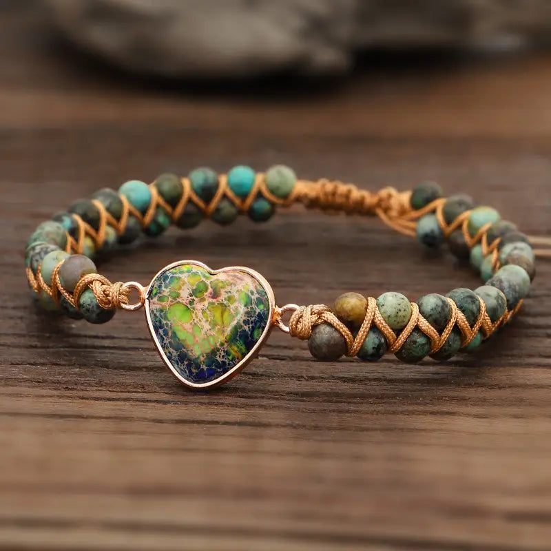Handmade woven heart stone pendant bracelet featuring a heart-shaped turquoise and green stone pendant with a gold-tone metal frame on a braided cord with mixed teal and earth-toned beads.