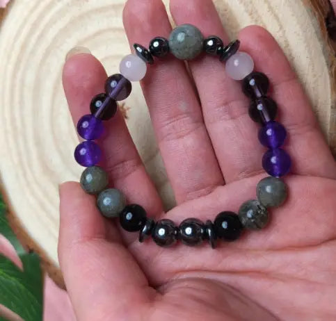A hand holds a natural amethyst and magnetic stone beaded unisex bracelet featuring alternating round beads of deep purple amethyst, light pink quartz, dark gray smoky quartz, and glossy black hematite.