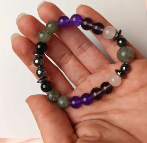 A hand holds a natural amethyst and magnetic stone beaded unisex bracelet featuring alternating purple, black, greenish-gray, and translucent white round beads.