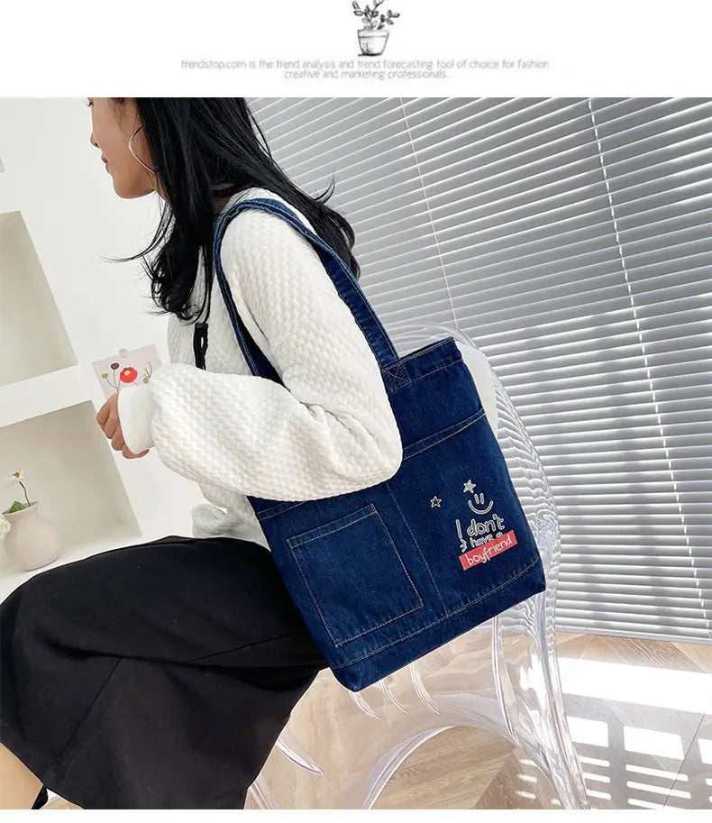 Blue denim embroidered tote bag with a smiling face and star design on the front pocket and a soft handle.