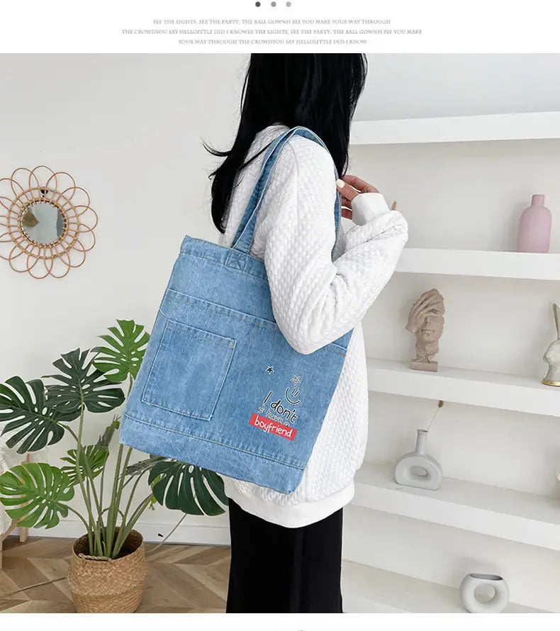 Light blue denim tote bag with a front pocket featuring embroidered text and a red rectangular patch with a soft handle.