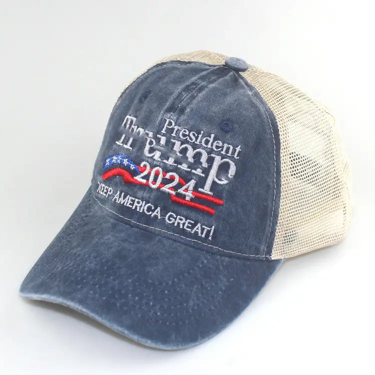 A faded blue and beige denim trump 2024 embroidered trucker hat with red stripes stars and the slogan keep america great on the distressed front panel.