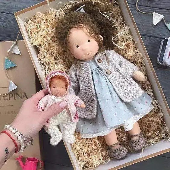 Eine Hand hält eine handgemachte Plüschbaby-Puppe mit goldenen lockigen Haaren, gekleidet in ein sanftes Rosa und mit weißem handgestricktem Blumendetail, mit einer Größe von 30 cm.