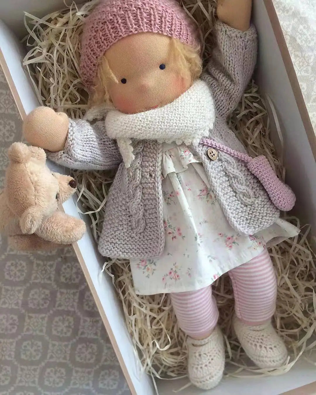Handgemachte Plüschpuppe mit goldenen lockigen Haaren und einem weich handgestrickten Design, mit einer rosa Strickmütze, einem hellgrauen Zopfmantel, einem Blumenkleid und gestreiften Leggings, Größe 30 cm.