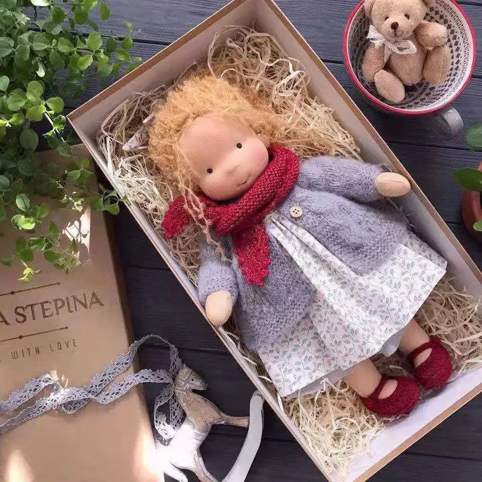 Handgemachte Plüschpuppe mit goldenen lockigen Haaren, mit einem grauen Strickmantel über einem weißen Blumenkleid und einem roten Stricksal, Größe 30 cm.