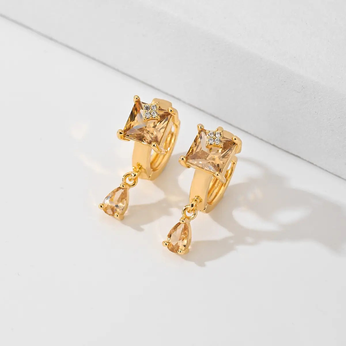 Des boucles d’oreilles créoles goutte en pierre précieuse géométrique élégantes en ton or, présentant une pierre ambrée à taille carrée, un petit accent de type diamant et un pendentif ambré en forme de goutte.
