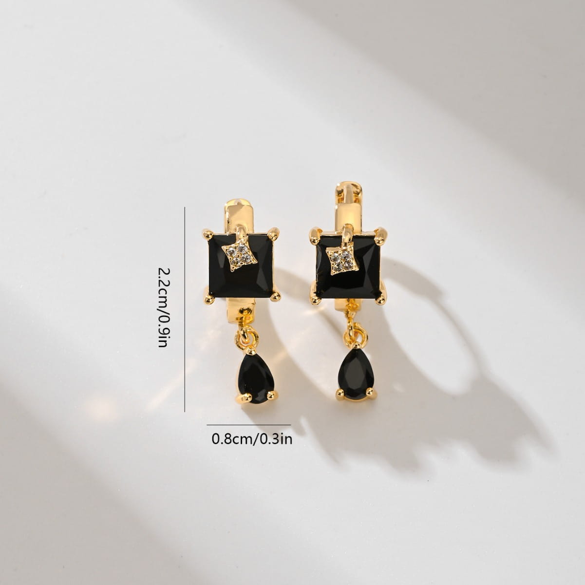 Des boucles d’oreilles créoles goutte en pierre précieuse géométriques élégantes en ton or, présentant une pierre noire en onyx à taille carrée avec un petit accent de diamant au-dessus d’une pierre noire en forme de goutte.