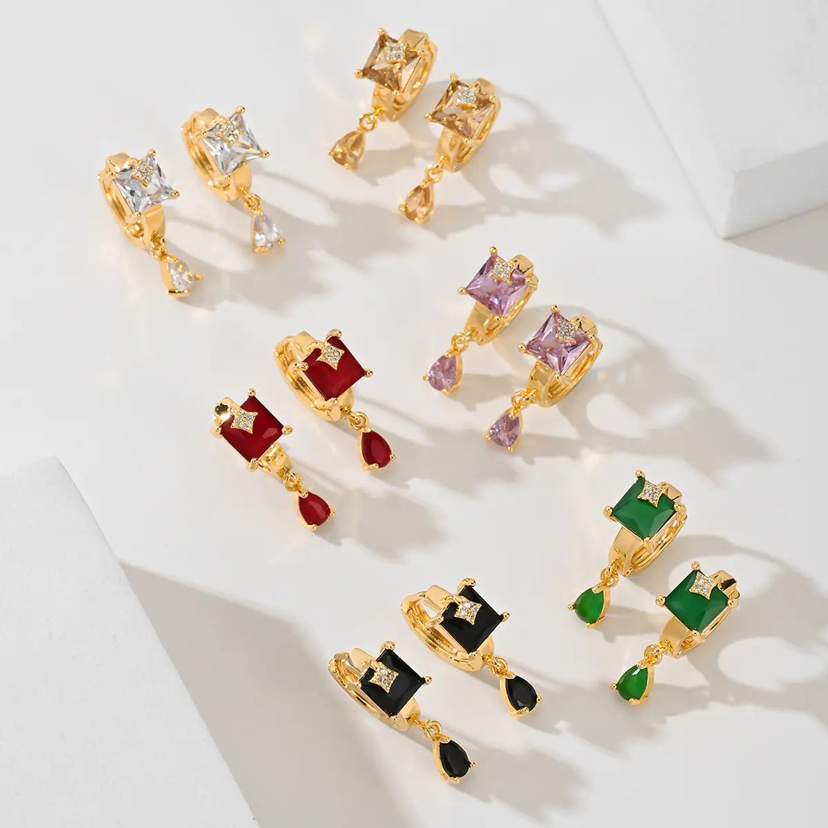 Des boucles d’oreilles créoles goutte en pierre précieuse géométriques élégantes, avec des clous en métal doré et des pierres précieuses colorées à taille carrée, agrémentées de pendants transparents en forme de goutte, aux tons violet, rouge, noir et vert.
