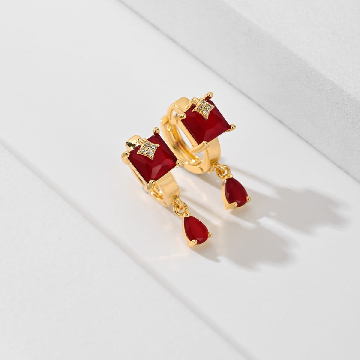 Des boucles d’oreilles créoles goutte en pierre précieuse géométriques élégantes en ton or, présentant des pierres carrées et en forme de goutte rouges, rehaussées de petites pierres au rendu de type diamant.