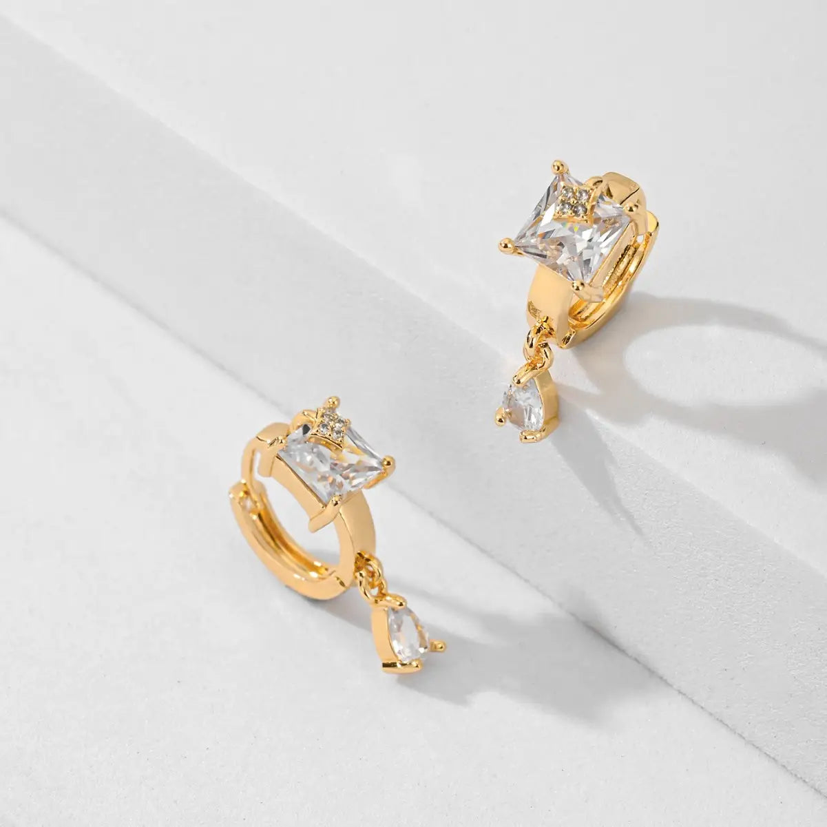 Des boucles d’oreilles créoles goutte en pierre précieuse géométriques élégantes en ton or, présentant un cristal clair à coupe carrée en partie supérieure et un cristal en forme de goutte suspendu, rehaussé de minuscules pierres étincelantes.