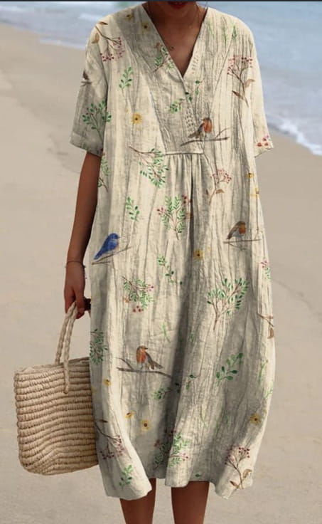 A soft off-white linen floral print v neck loose maxi dress featuring a delicate print of birds and foliage in muted greens blues and yellows.