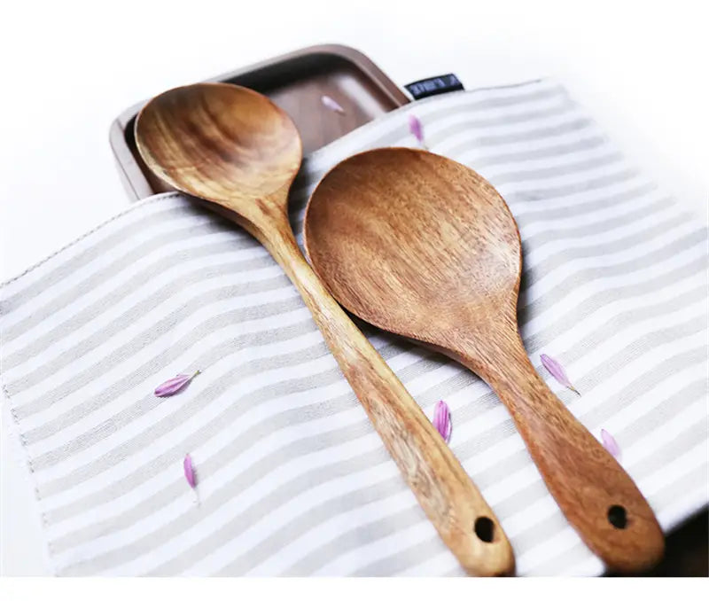 Dos cucharas de cocina de madera de teca con tonos naturales, con superficies pulidas y lisas y mangos redondeados, que presentan pequeños orificios en los extremos, incluidas en un set de utensilios de cocina de cuatro piezas.