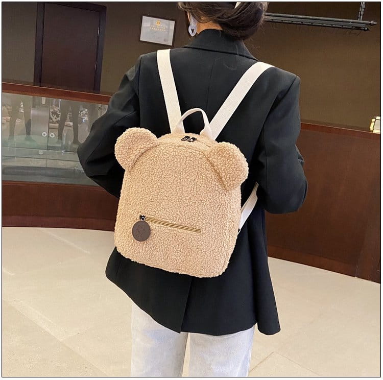 beige plush teddy bear shaped backpack with white straps and a round brown zipper pull made of fuzzy fleece for a cute and cozy rucksack option.