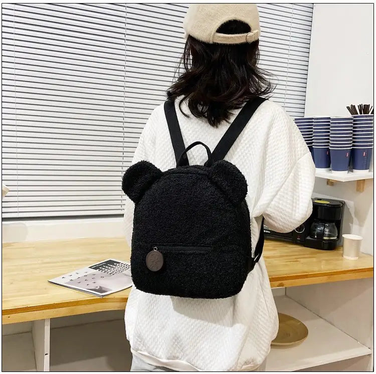 Black fuzzy bear shaped plush backpack with fleece material, plush ears, and a round metallic charm on the front pocket.