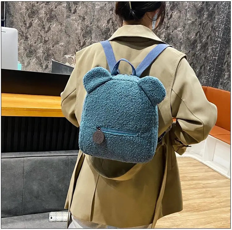 Blue fuzzy bear-shaped backpack made of soft fleece material with matching blue straps shown from the back side.