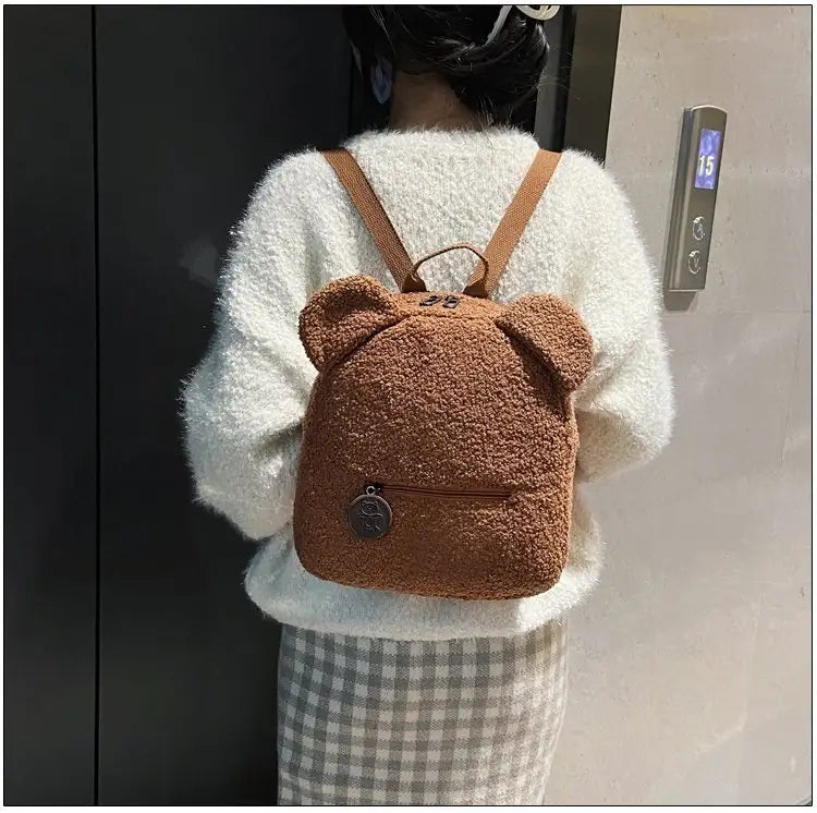 brown fuzzy bear-shaped backpack made of plush fleece with tan straps and a small circular metal charm on the front pocket designed as a cute rucksack.